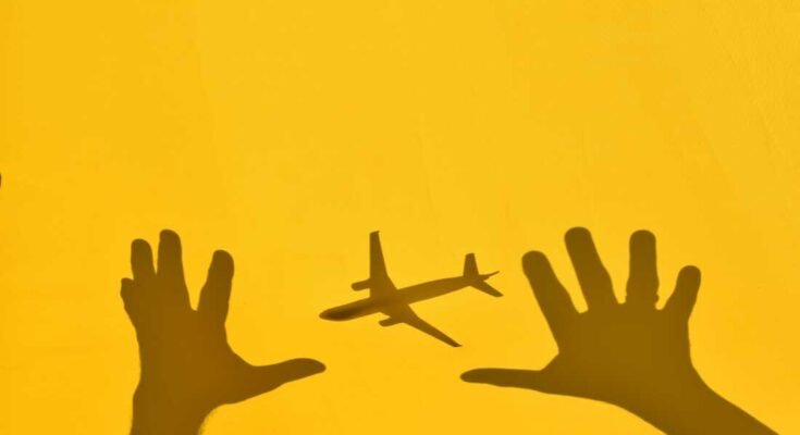 Fotografía de una sombra de un avión y dos personas duras que llegan al avión, contra una pared amarilla.