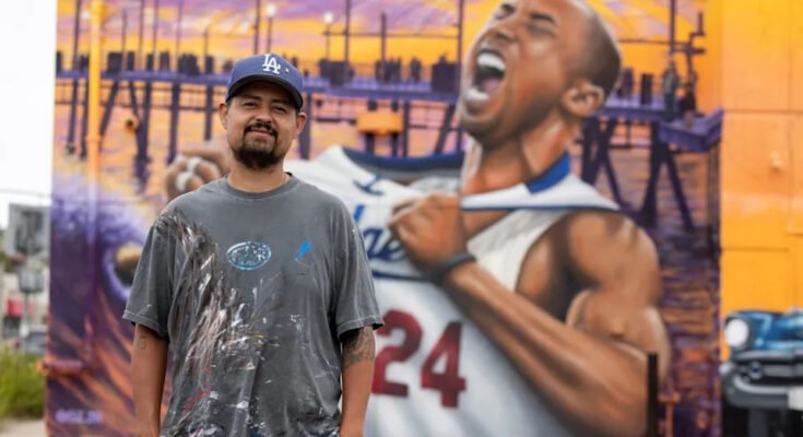 Un hombre con una gorra de los Dodgers y una camiseta negra desteñida se encuentra en sus bolsillos frente a una pared de Kobe Bruy